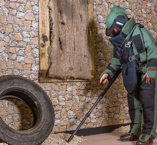 A Kapri Corp EOD technician in a full bomb-disposal suit probing a suspect device with a long-reach tool